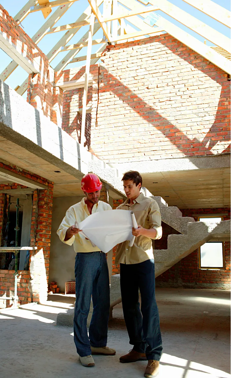 Two construction workers, known as top Los Angeles custom home builders, review blueprints inside a partially built brick structure with exposed beams.