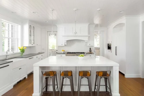 Bright White Kitchen With A Large Island, Wooden Stools, Hanging Lights, And Stainless Steel Appliances, Offering A Sleek Look That Complements Any Home Renovation Project, Including Bathroom Remodeling.