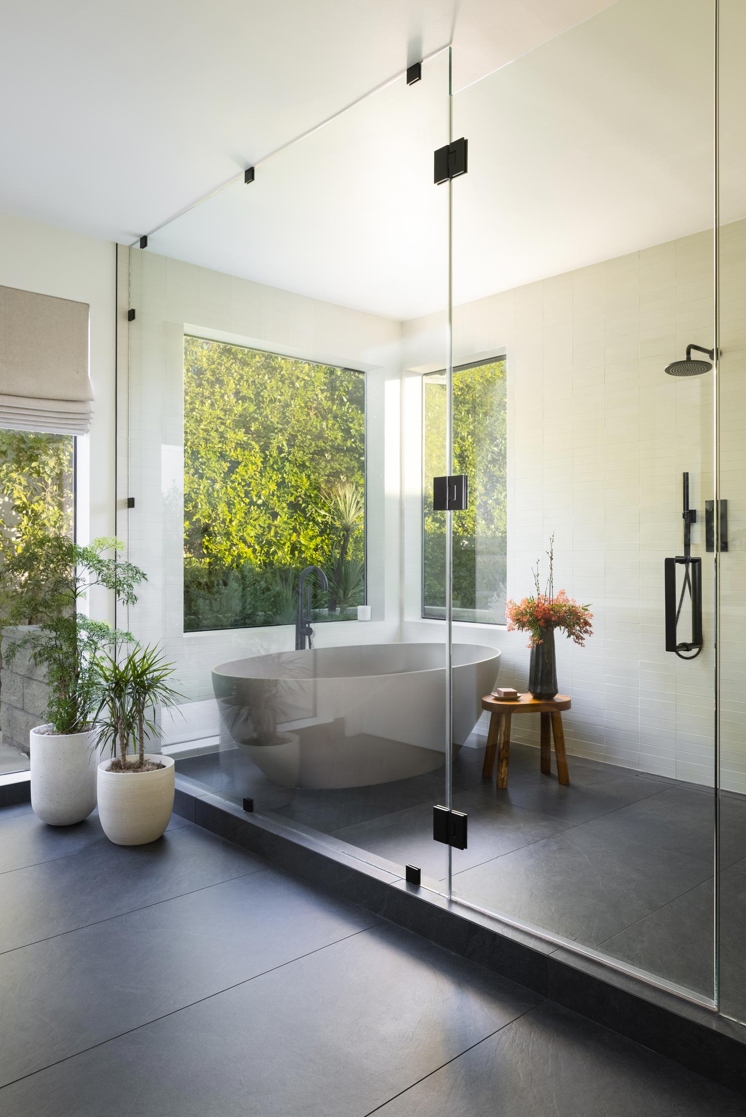 Modern bathroom with large bathtub, glass-enclosed shower, plants, and greenery visible through windows.