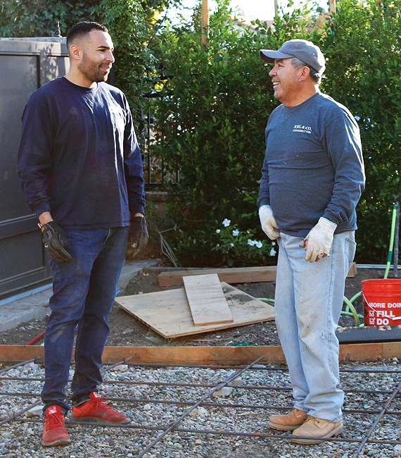 Our-approach About two men in work clothes and gloves chat and smile at a construction site with tools and boards around them.