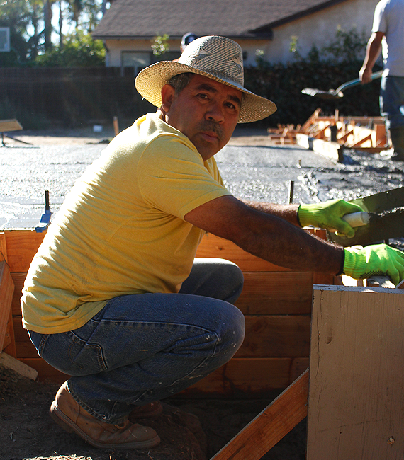 Raul-portfolio About: Man wearing a straw hat and yellow shirt building a wooden frame at a construction site outdoors.