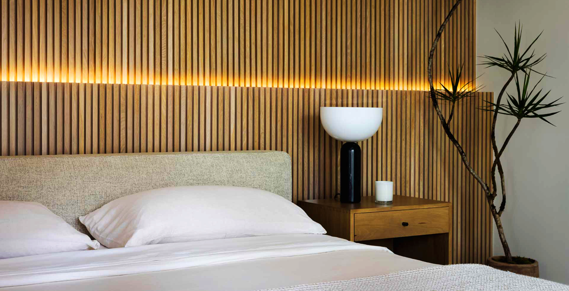 About this space: This minimalist bedroom features a stunning wood slat wall, soft bedding, and a stylish bedside table topped with a lamp and candle. A tall plant completes the serene atmosphere, making it the perfect retreat for relaxation.
