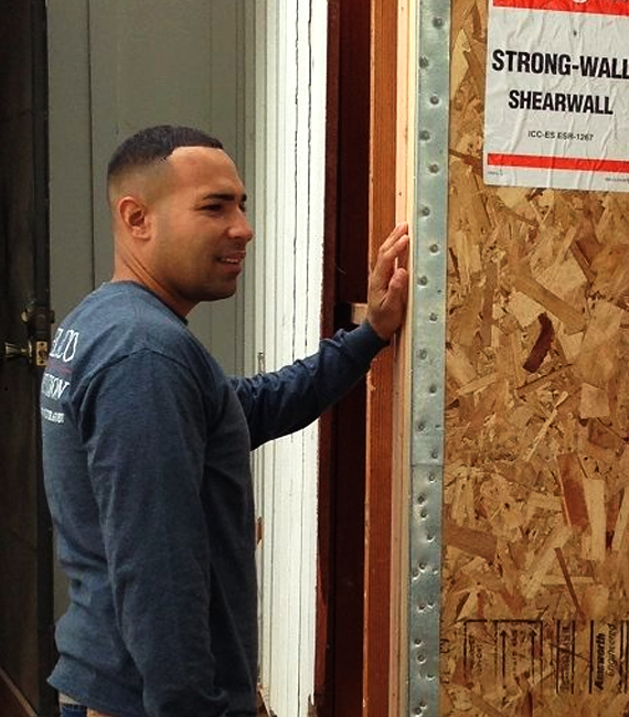joel-portfolio About: Man standing by a wooden wall under construction, touching the frame near a “Strong-Wall Shearwall” sign.